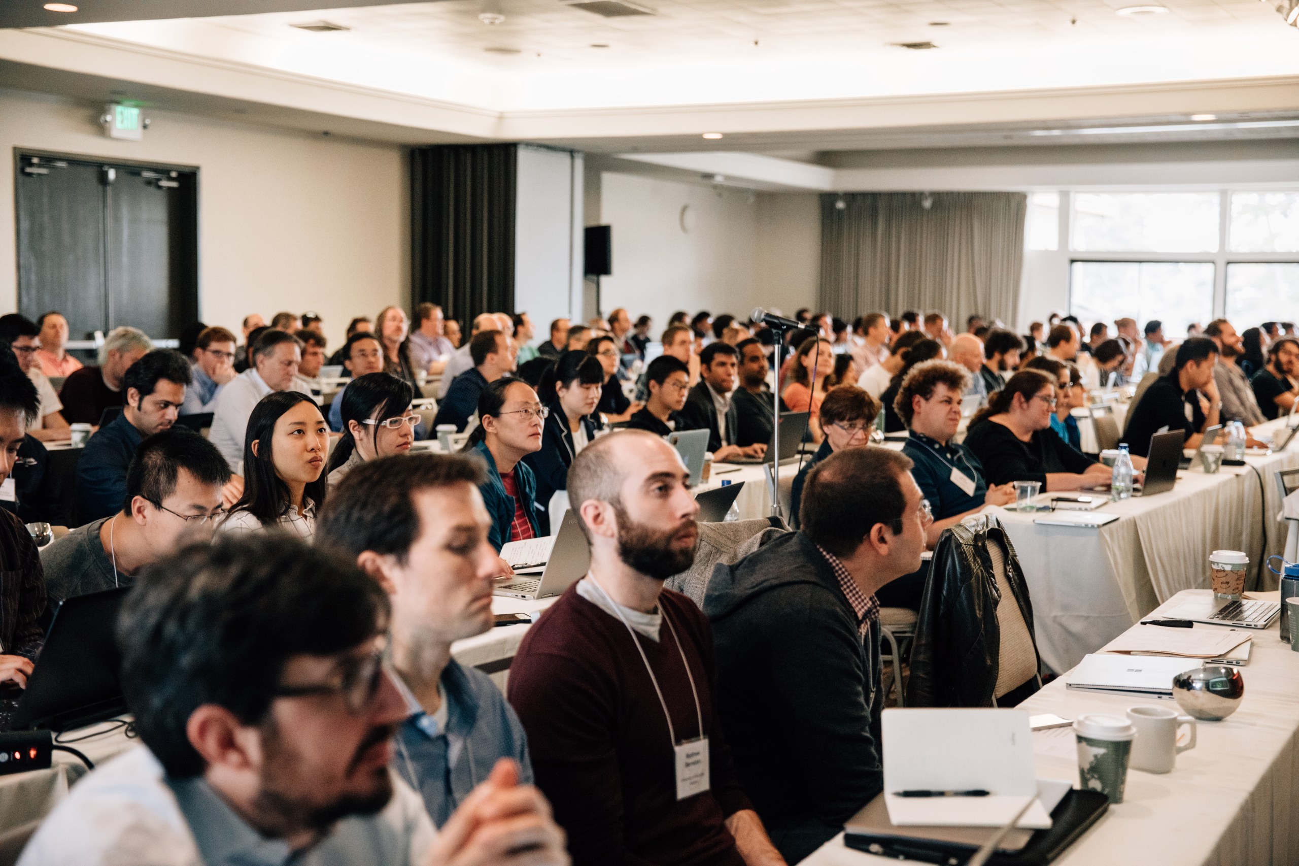 A large group of people sitting at tables in a conference hall. Some individuals are using laptops, while others are interacting with one another.