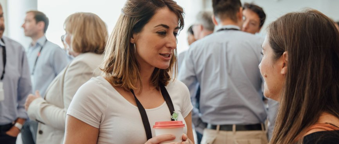Two people standing in a busy room, engaged in conversation; other people are visible in the background.