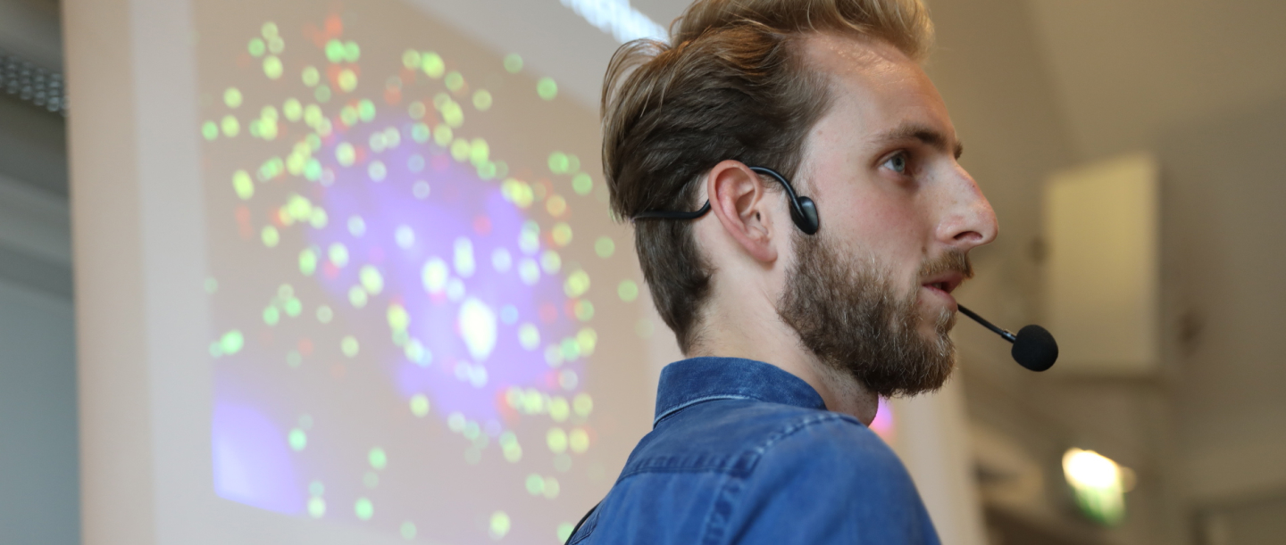 Person wearing a headset microphone, speaking in front of a screen displaying a colorful data visualization.