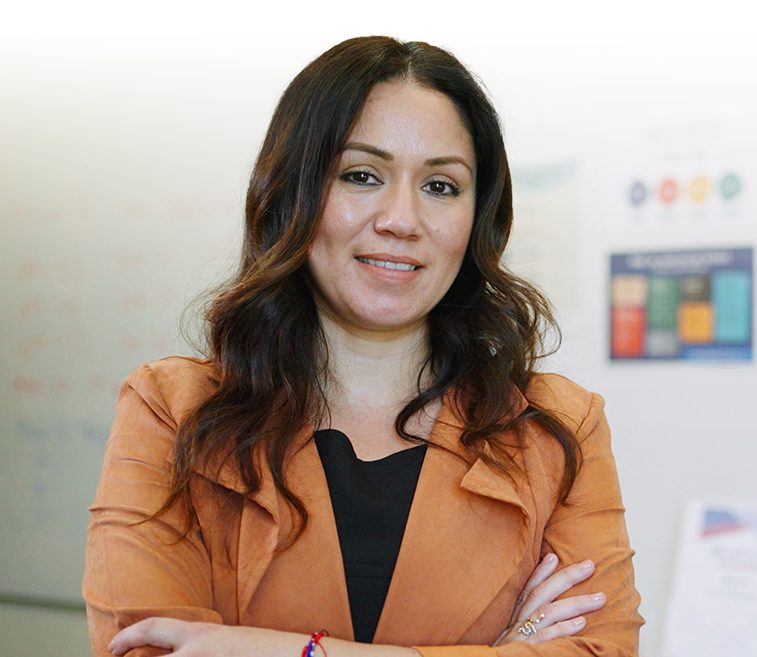 Woman with brown, wavy hair wearing a blazer with her arms crossed and standing in a classroom.