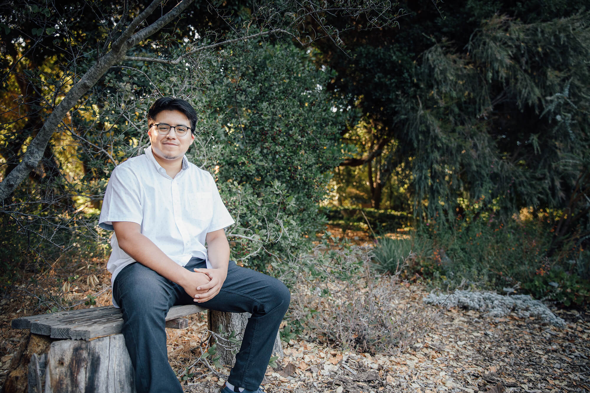 A man in a white shirt and jeans sits and has trees behind him.