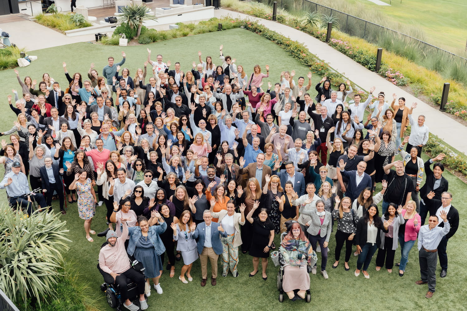 Meeting participants gather close together to smile and wave at the camera. They pose together outside on a grassy lawn.