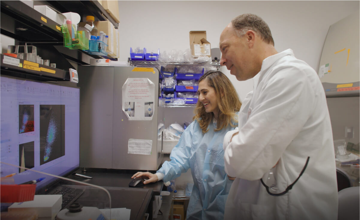 Two scientists smile while standing in a lab setting and looking at a computer screen.