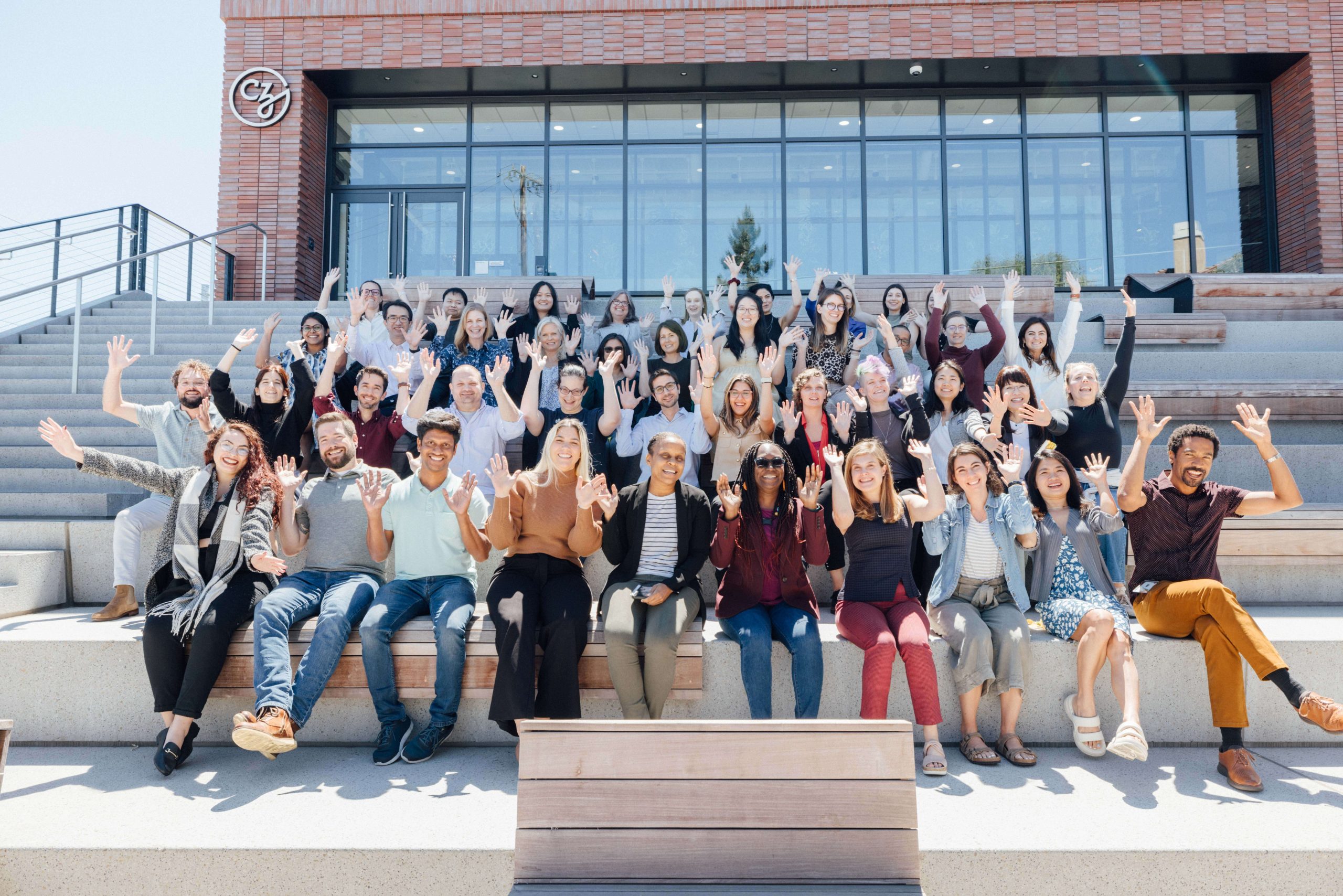Participants of the CZI Reproductive Tissue Atlas Annotation Jamboree sit on the steps in front of the CZI headquarters in Redwood City, CA, and wave at the camera.