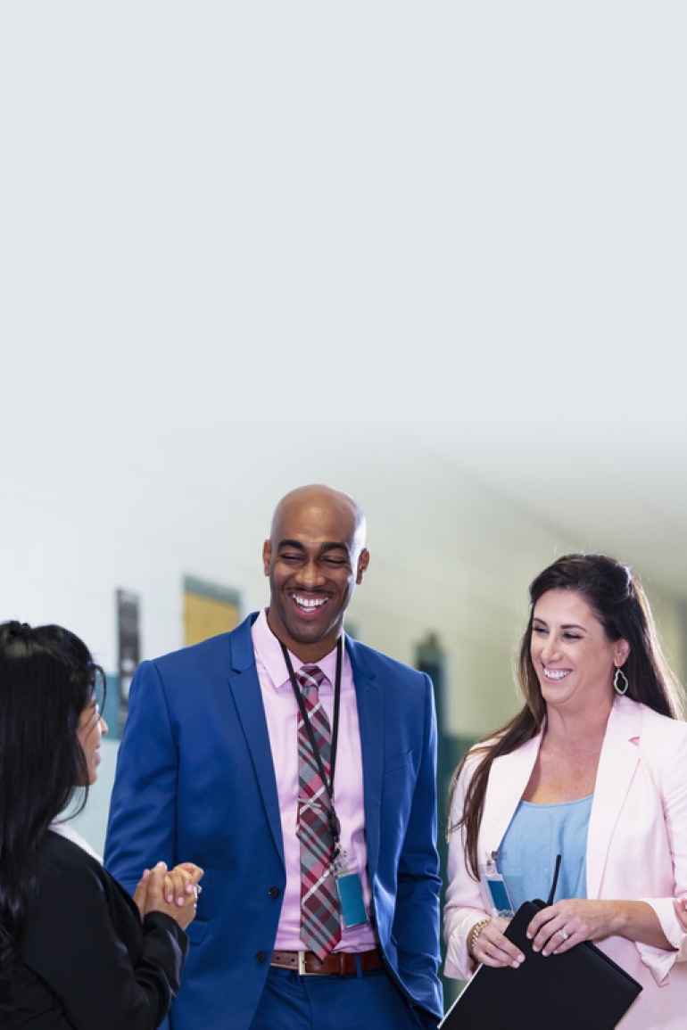 A diverse group of educators stand in a school hallway, engaged in a friendly discussion.