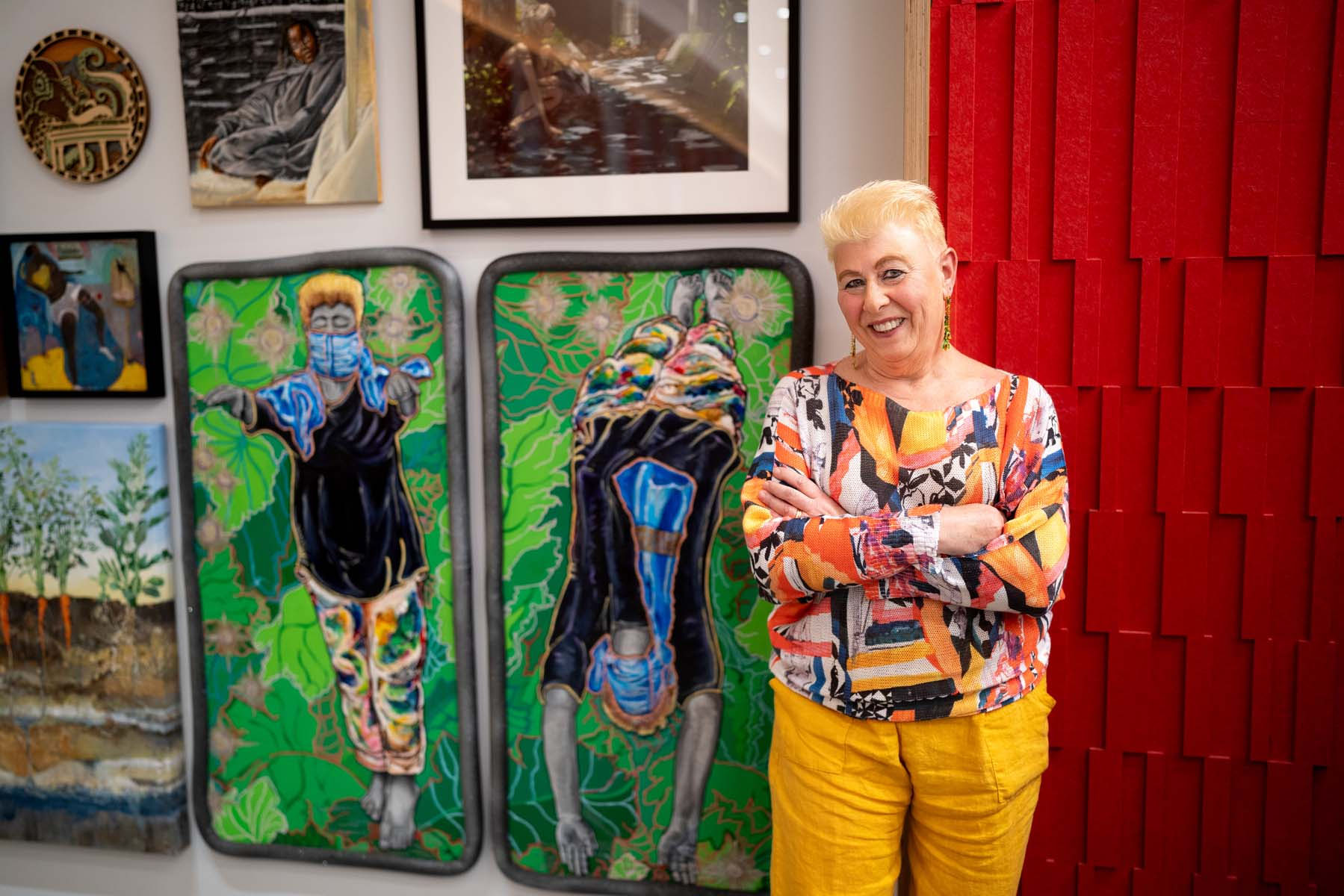 A person stands in front of their artwork as they smile at the camera with their arms crossed over their chest.